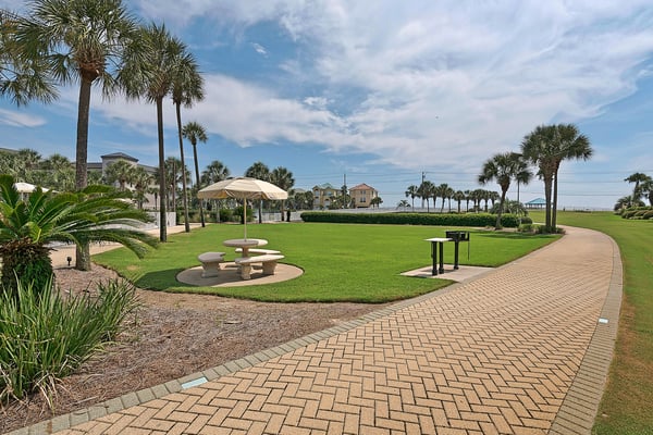 A spacious outdoor recreation area features a picnic table with umbrella shade, surrounded by manicured lawns and swaying palm trees, offering a tranquil spot for relaxing or dining alfresco.
