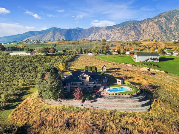 This aerial view showcases the property nestled in a scenic valley surrounded by mountains, orchards, and farmland, with a private pool and terraced landscaping.