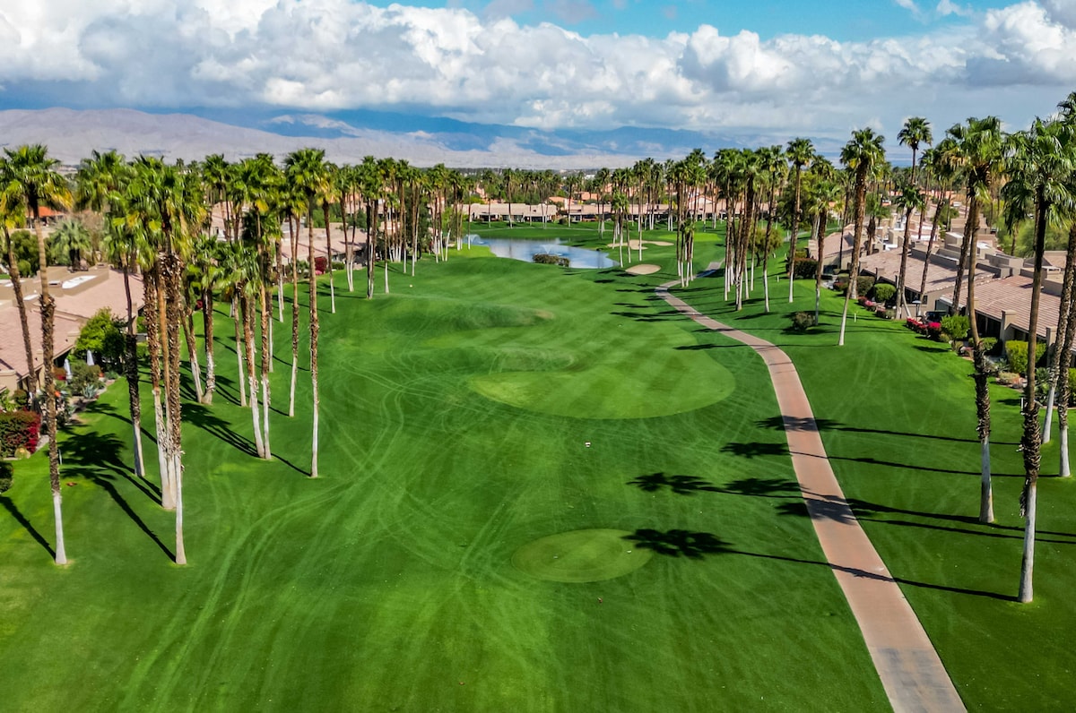 An aerial view captures the pristine golf course, framed by towering palms and a serene water feature, with residential buildings and mountain vistas in the distance.