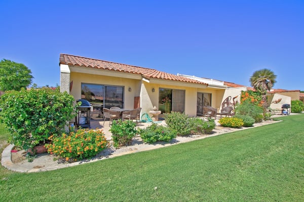 A warm desert retreat with Spanish tile roof, lush green lawn, and colorful flower beds welcoming you to sunny outdoor living and relaxation under clear blue skies.