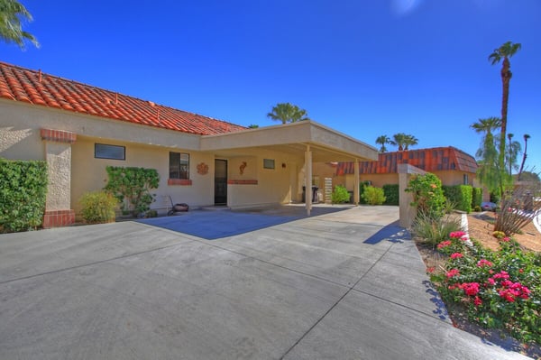 This desert retreat features a welcoming covered entrance with charming terracotta tile roofing, surrounded by palm trees and vibrant flowering plants that frame your private oasis.