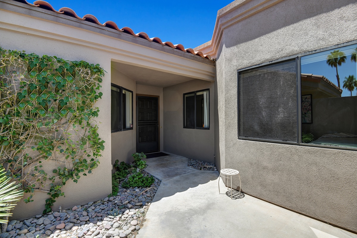 The property features a charming stucco exterior with terra cotta tile roofing and a welcoming entrance framed by lush greenery—a perfect desert retreat in a quiet neighborhood.