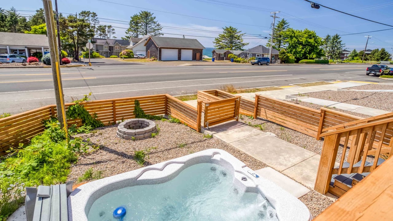 The property's outdoor area features a private hot tub, fire pit, and wood-fenced patio positioned along a residential street. Nearby houses and natural scenery characterize the neighborhood.