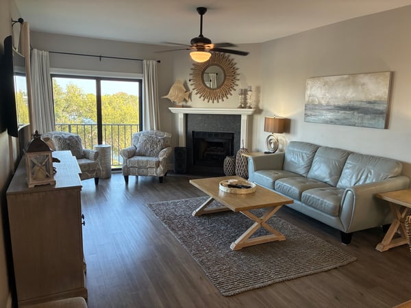 Sink into the plush leather sofa and let your cares drift away in this inviting living space, where natural light floods through glass doors framing peaceful treetop views and a cozy fireplace awaits your evenings.