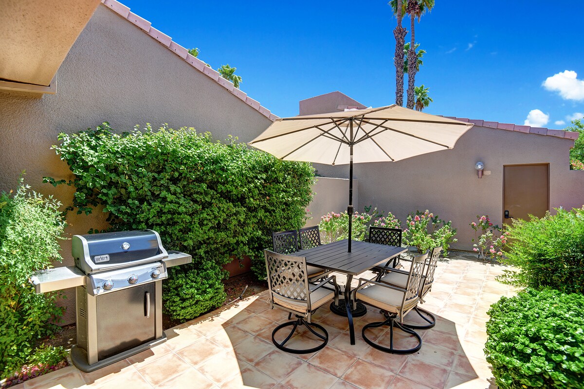 Gather around your private outdoor dining area, shaded by a market umbrella and surrounded by lush greenery, with a gleaming grill ready for your cookouts under sunny skies.