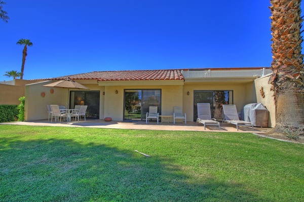 The property sits on a well-maintained lot with palm trees, a red tile roof, and a private patio area bordered by lush grass.