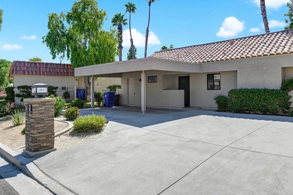 Single-story stucco residence featuring a covered carport and manicured desert landscaping with palm trees under clear blue skies.