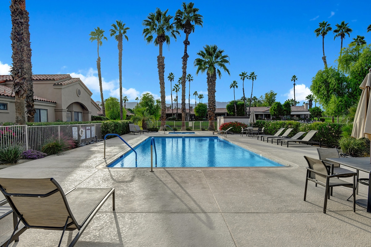 The community pool surrounded by towering palms invites sun-soaked relaxation on warm desert days. Lounge chairs await under clear blue skies, while mountains frame the horizon beyond the lush greenery.