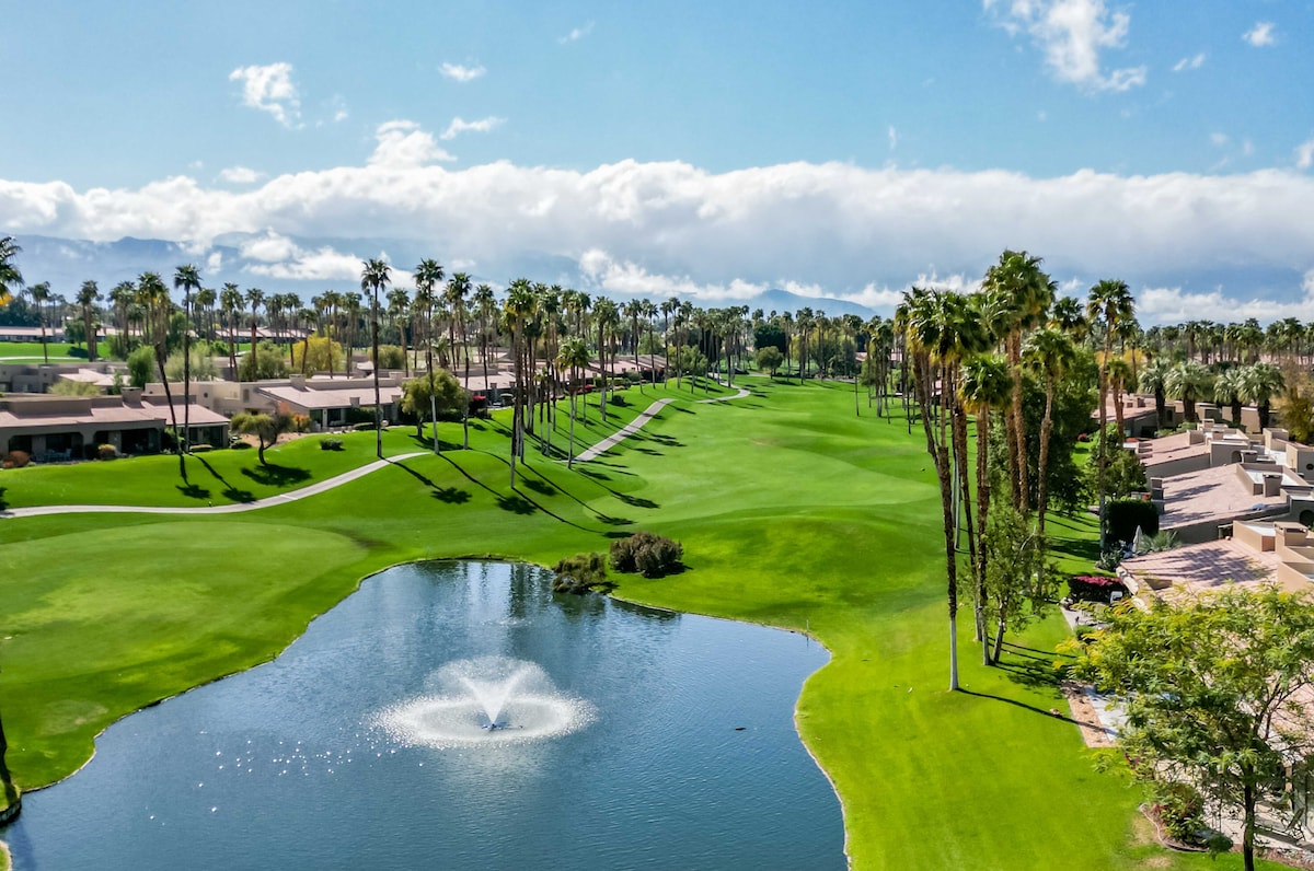An aerial view captures the resort's lush golf course winding through tall palm trees and neighboring residential buildings under a bright desert sky.