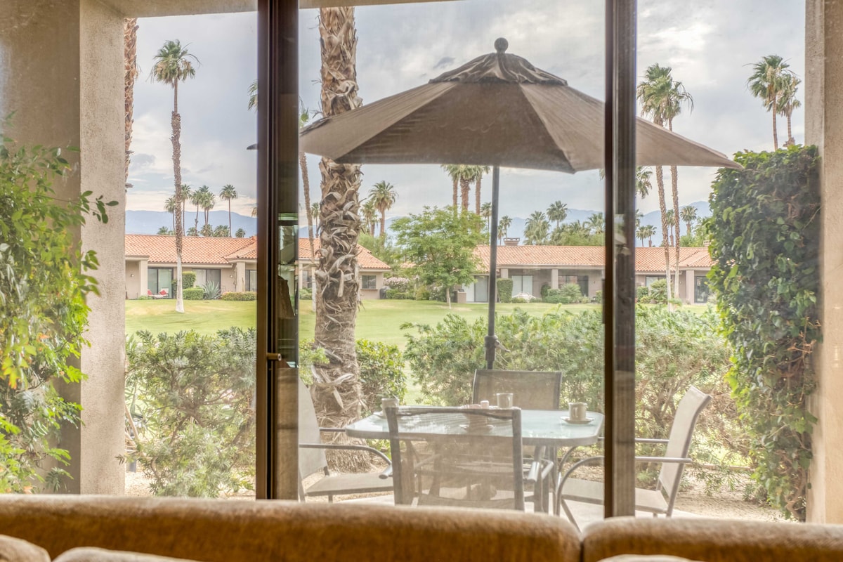 Step through the sliding glass door onto your private patio, where palm trees sway above a grassy courtyard and mountains rise in the distance—your peaceful desert retreat awaits.