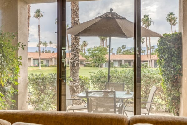 Step through the sliding glass door onto your private patio, where palm trees sway above a grassy courtyard and mountains rise in the distance—your peaceful desert retreat awaits.