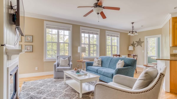 Unwind in your cozy living room, where a plush blue sofa and soft armchairs invite you to relax beneath the gentle breeze of ceiling fans. Sunlight streams through large windows, warming the hardwood floors and creating a peaceful retreat.