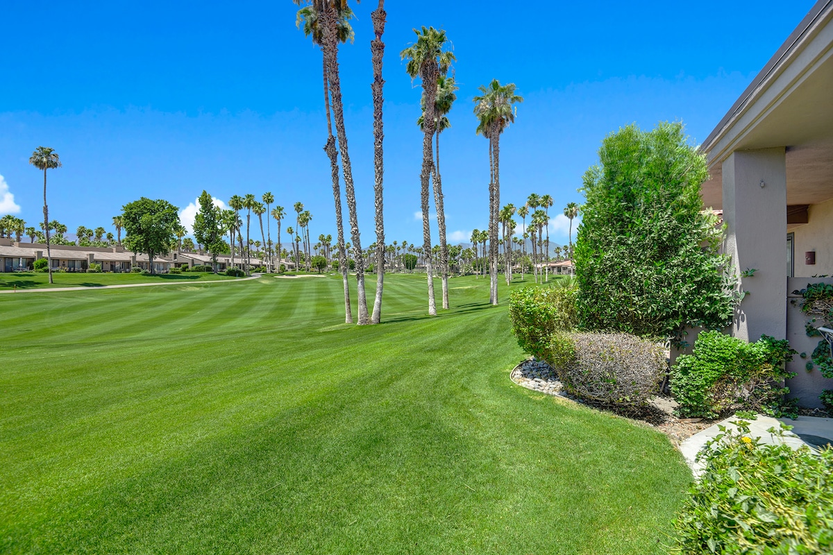A pristine fairway lined with towering palms stretches beyond your private patio, framed by lush greens and manicured landscaping under brilliant desert skies.