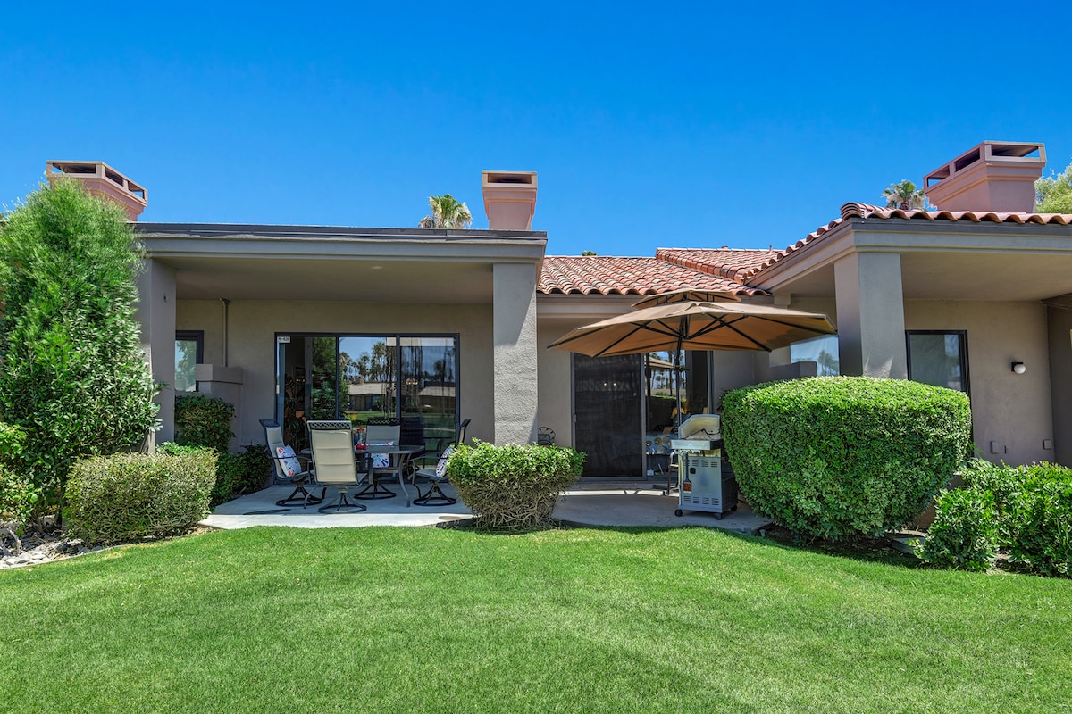 A charming desert retreat with covered patios and lush landscaping, set against a backdrop of palm trees and clear blue skies.