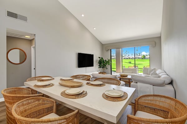 Unwind in your bright, open-concept living and dining area with soaring vaulted ceilings, natural light, and a sliding glass door that opens to lush green fairways and swaying palms.