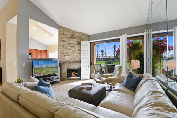Relax into plush seating while natural light streams through expansive glass doors, framing a view of the lush golf course beyond. The crackling fireplace adds warmth to your comfortable retreat.
